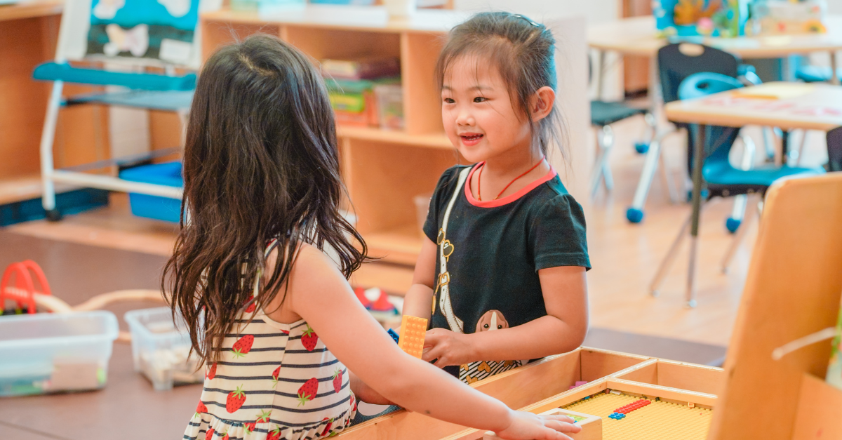 Two young children wearing dresses engage with each other while participating in an activity in a child care classroom.