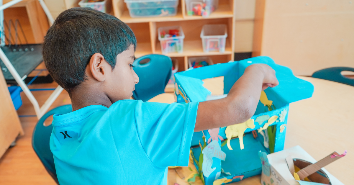 A child in a blue T-shirt works on an aquarium craft created with a cardboard box painted blue.