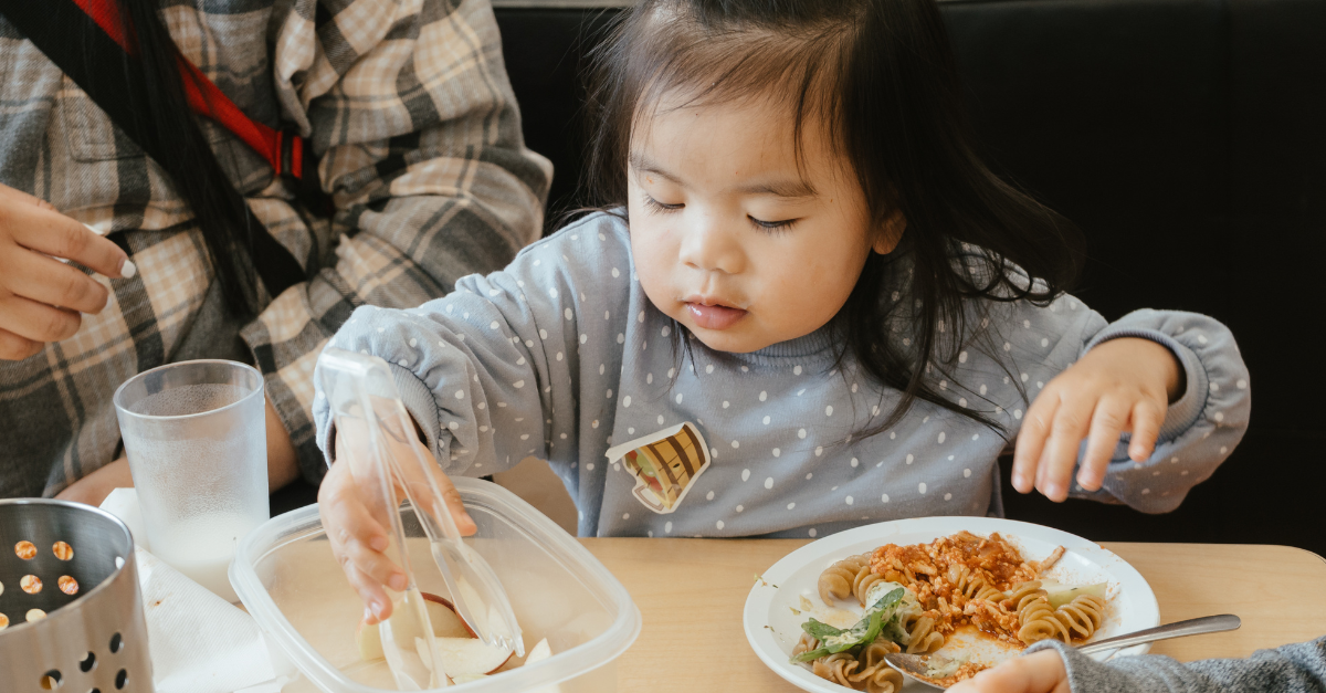 During lunchtime, a child uses child-safe plastic tongs to pick up an apple while eating pasta with sauce.