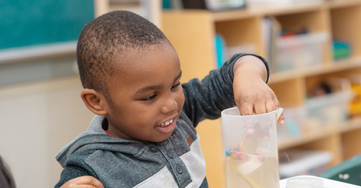 A child wearing a grey long-sleeve shirt with short hair smiles in awe while dipping a hand into a sensory water cylinder during a painting activity in a child care classroom.