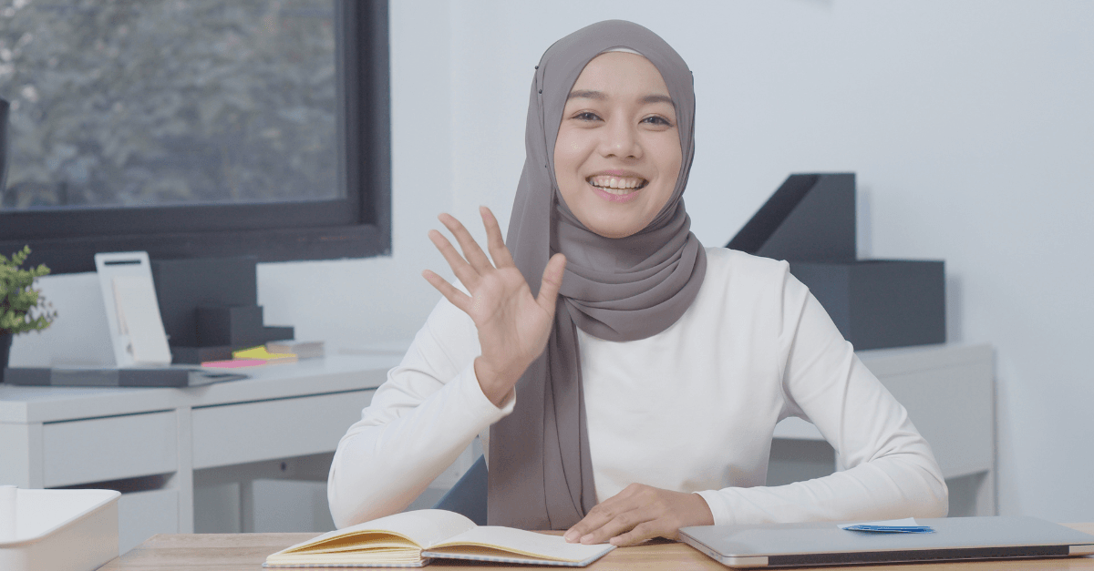 A person wearing a grey hijab and white long-sleeve shirt smiles and waves in a neutral office setting.