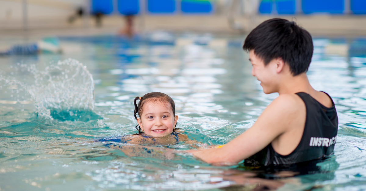 A smiling child practises swim skills one-on-one with an instructor at a Y M C A pool. The instructor wears a black swim top with the word "Instructor" on the back.